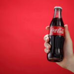 Coke’s 2027 Flavor Bombshell Hand holding a glass soda bottle against a red background